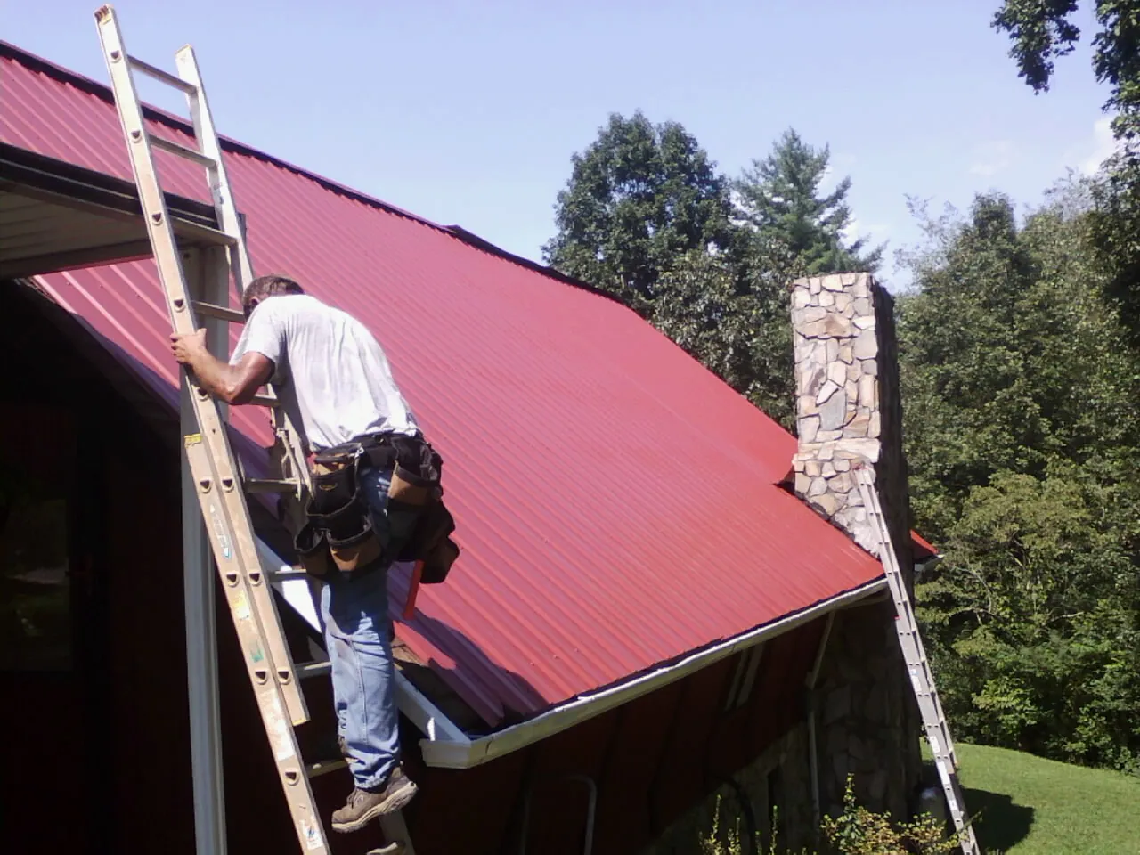 Roofer on a ladder beside a red metal roof and stone chimney — Soffit & Fascia Repair by Roofing Brothers in Walstonburg